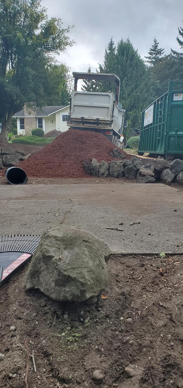 A truck unloading red mulch near a house and green dumpster; a large rock sits in the foreground.