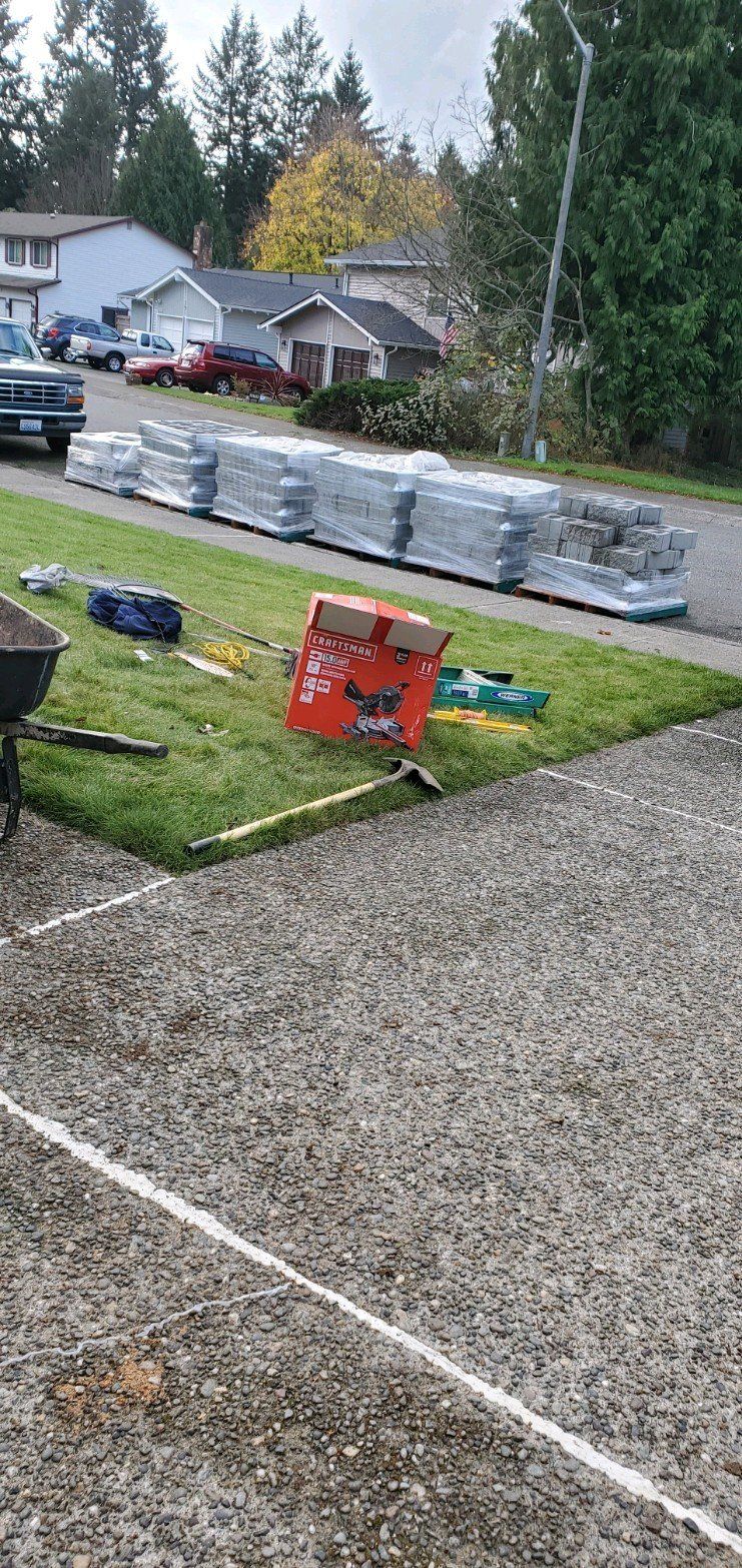 Construction materials stacked on a grassy area next to a gravel driveway in front of houses.