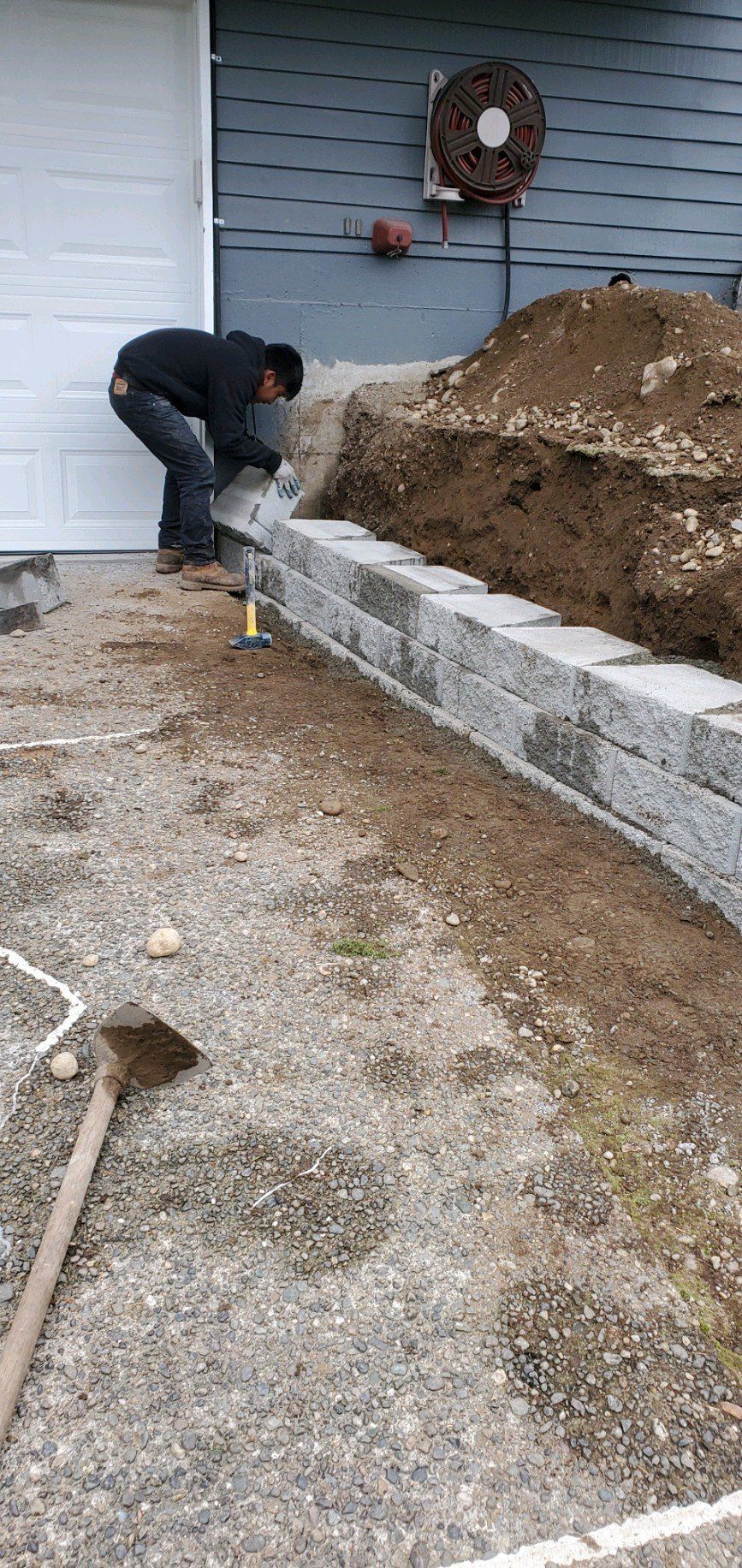 Person constructing a retaining wall outdoors. Gravel ground, dirt pile, blue siding.
