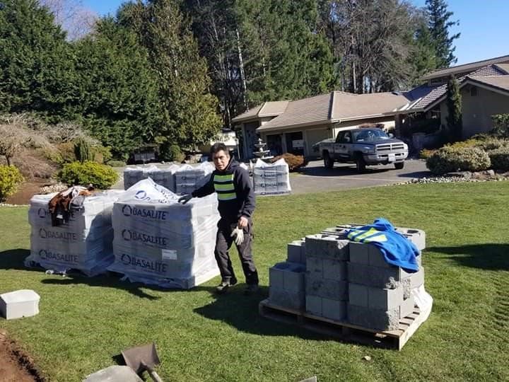 Man standing near stacks of gray blocks on a lawn with a house and truck in the background.