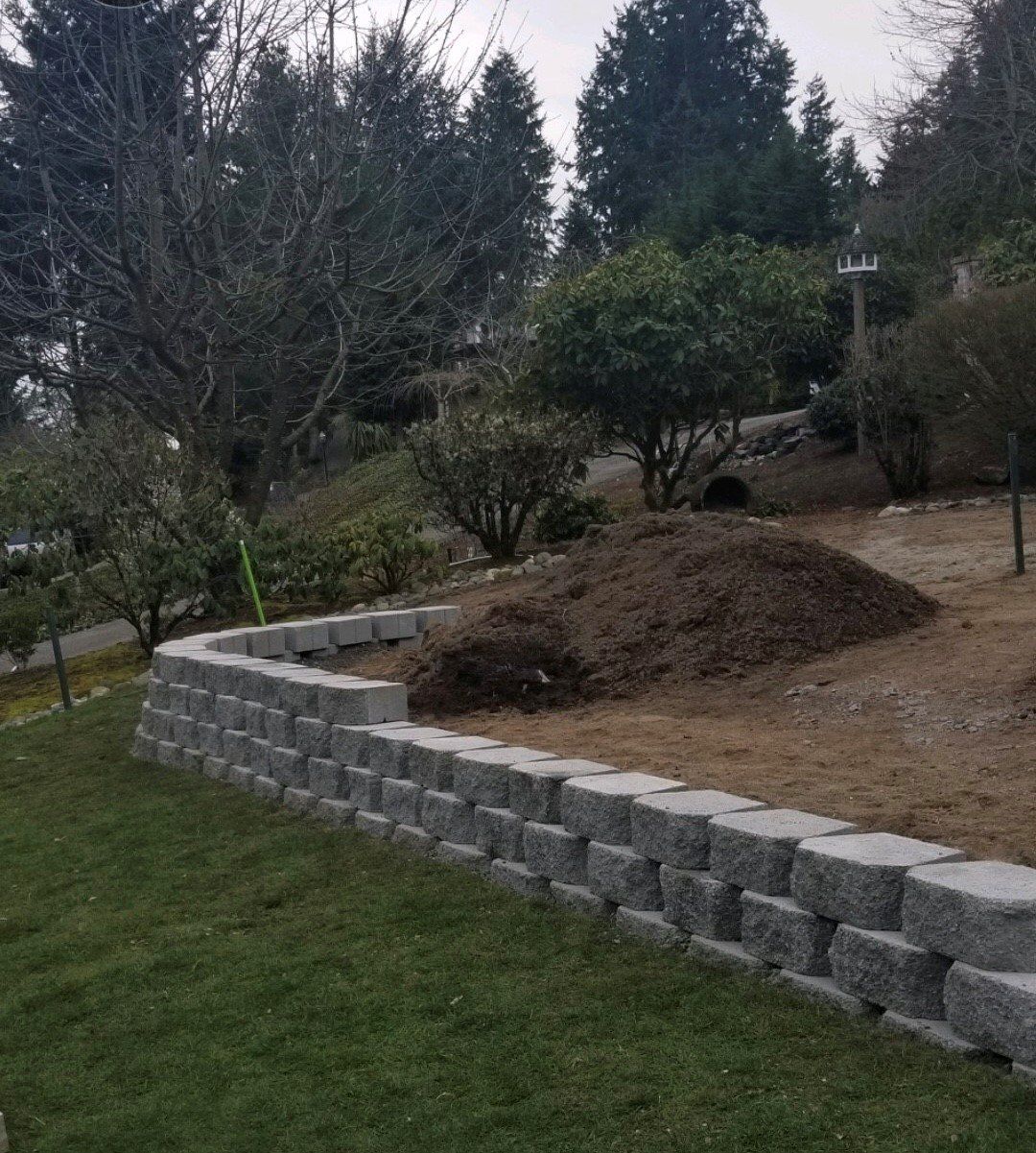 Gray block retaining wall on a grassy hillside, with mulch and trees in the background.