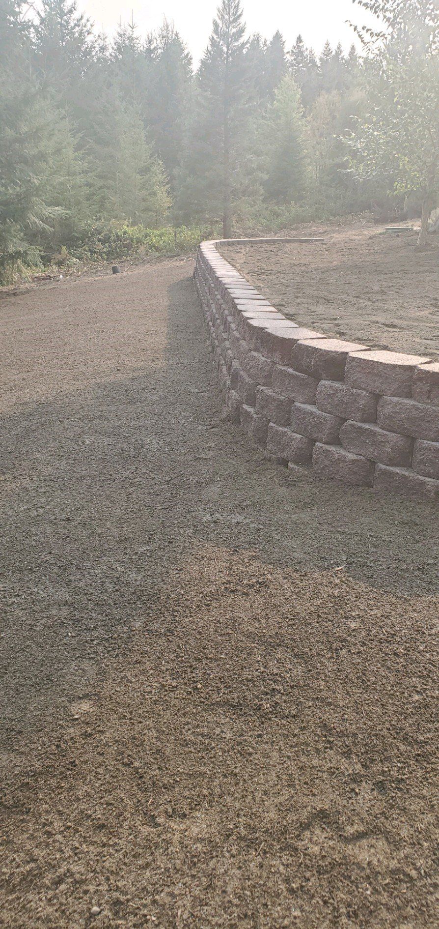 A low retaining wall made of stacked blocks curves along a gravel path. Trees are in the background.