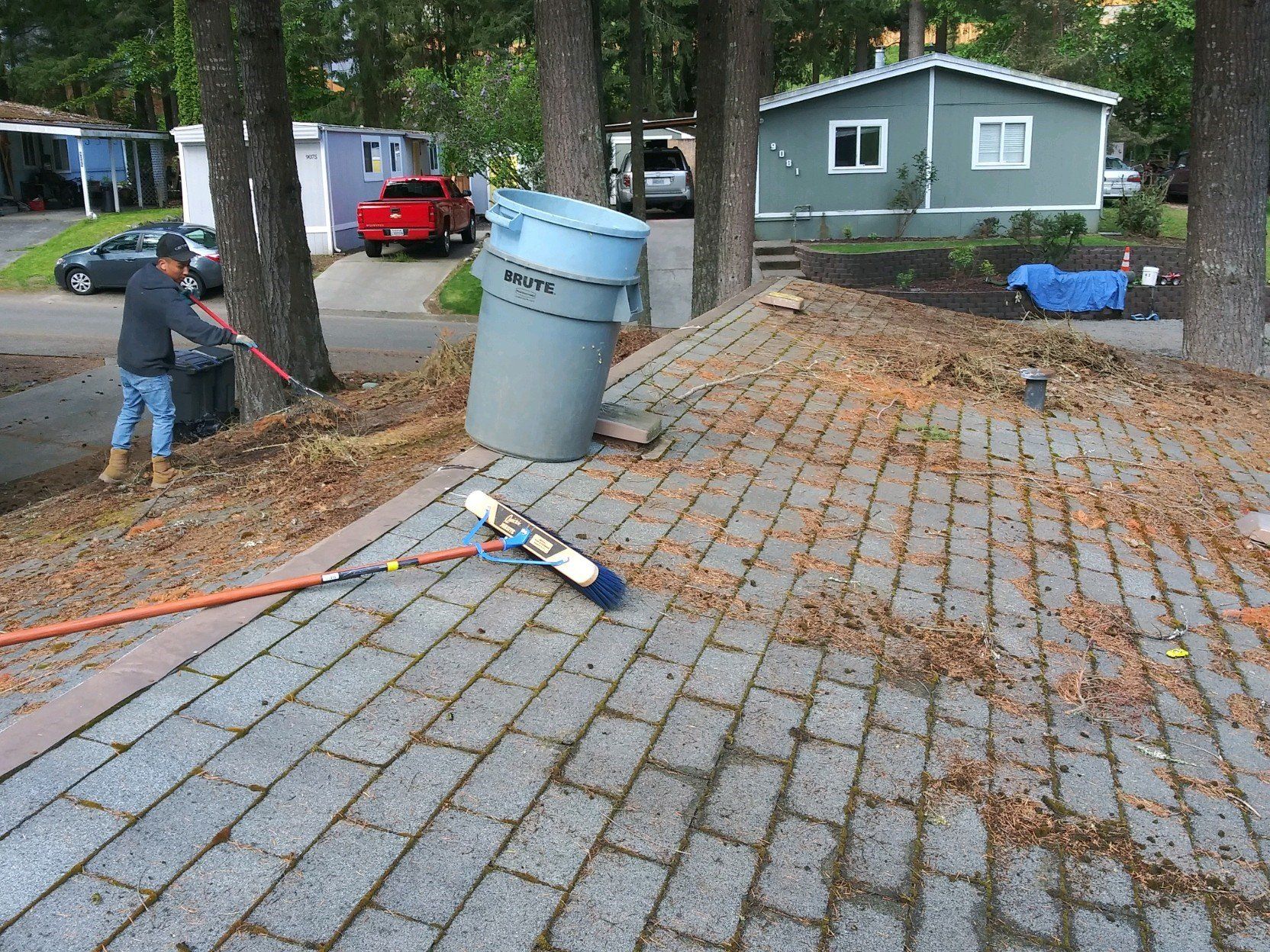 Man sweeping leaves off a roof, using trash can to collect debris, near trees and mobile homes.
