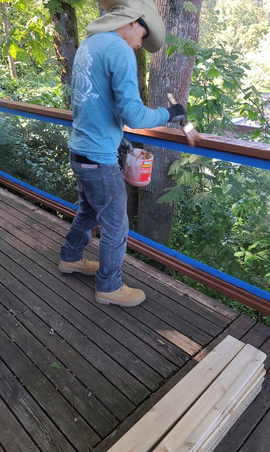 Person painting a deck railing outdoors with a paint bucket, surrounded by trees.