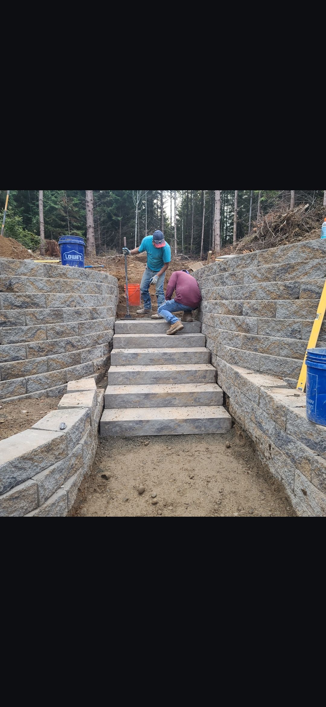 Workers building stone steps on a hillside. Two men are working with a bucket. Trees in the background.