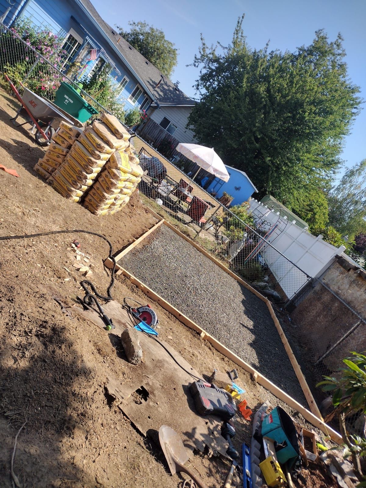 Backyard construction site; gravel inside wooden frame. Bags of cement stacked nearby, tools scattered.