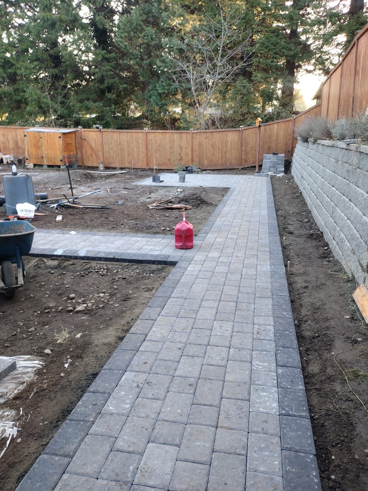 Brick pathway under construction; red propane tank in middle, next to retaining wall and wooden fence.
