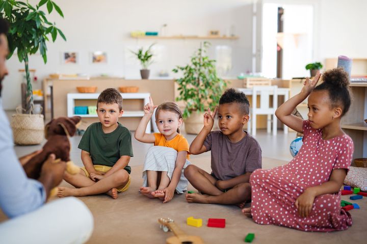 Children seated on floor with teacher; raising hands in a classroom.