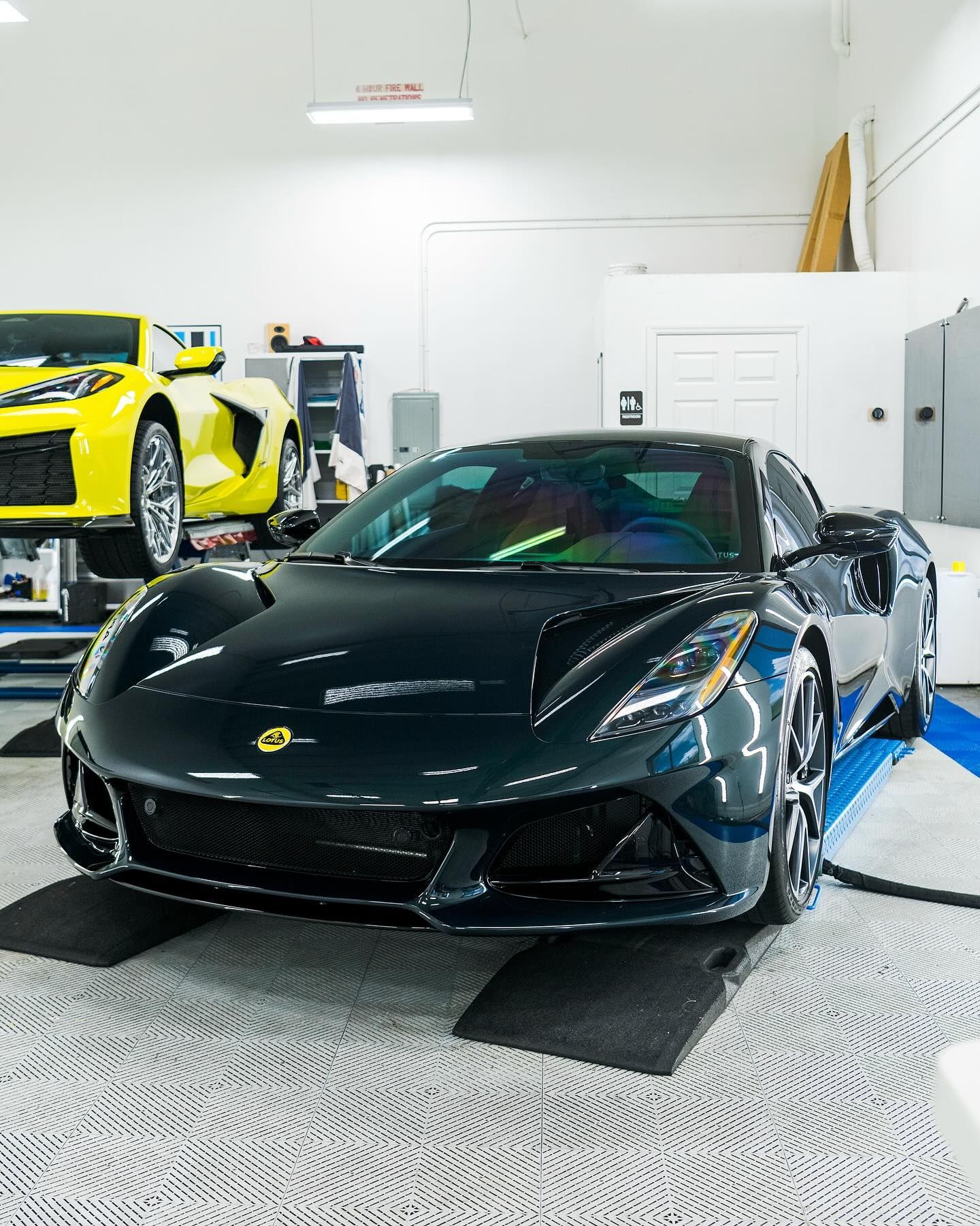 Black sports car in a bright garage, with a yellow car visible in the background.