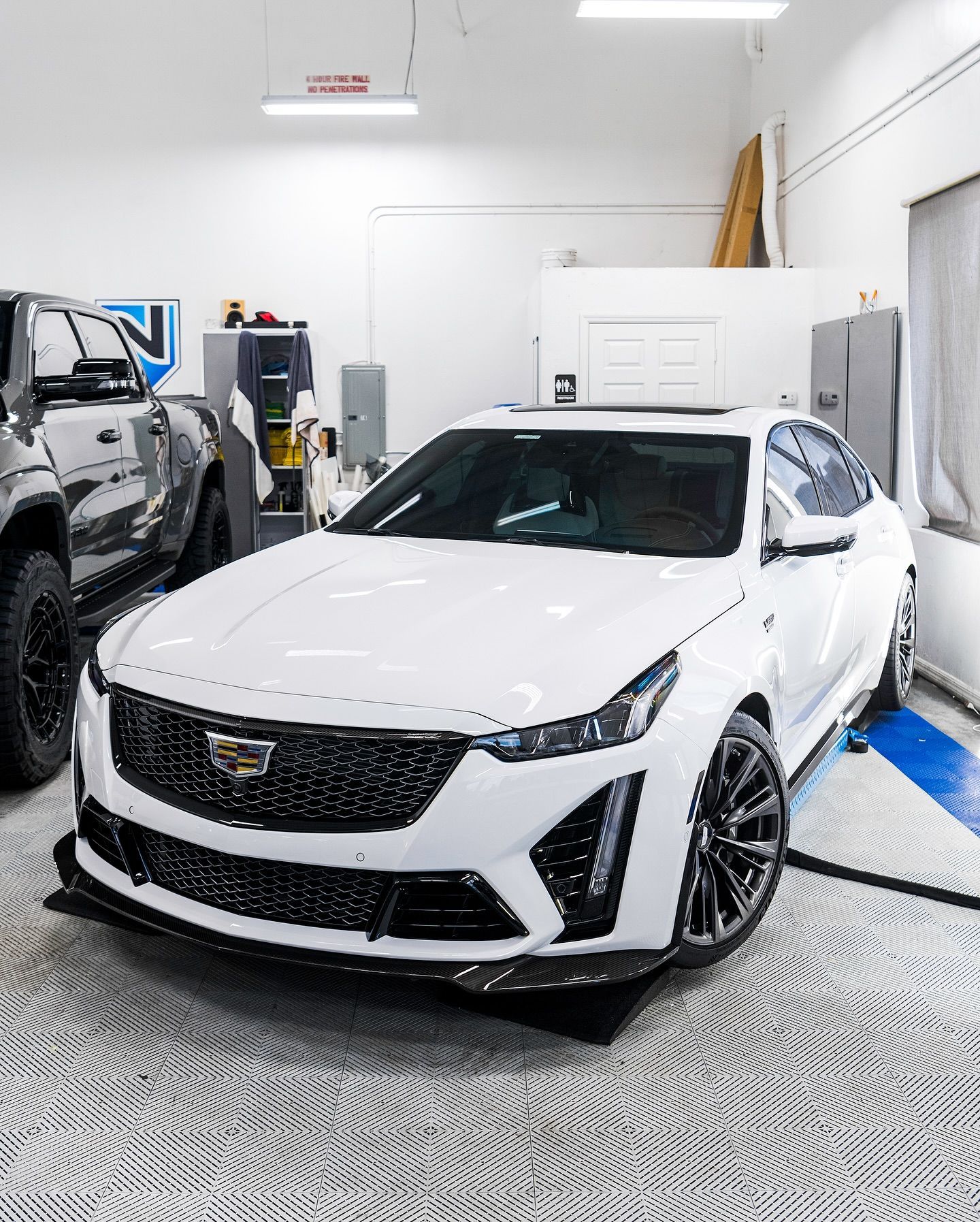 White Cadillac SUV in a garage, shown front-facing on a blue service lift next to another vehicle.
