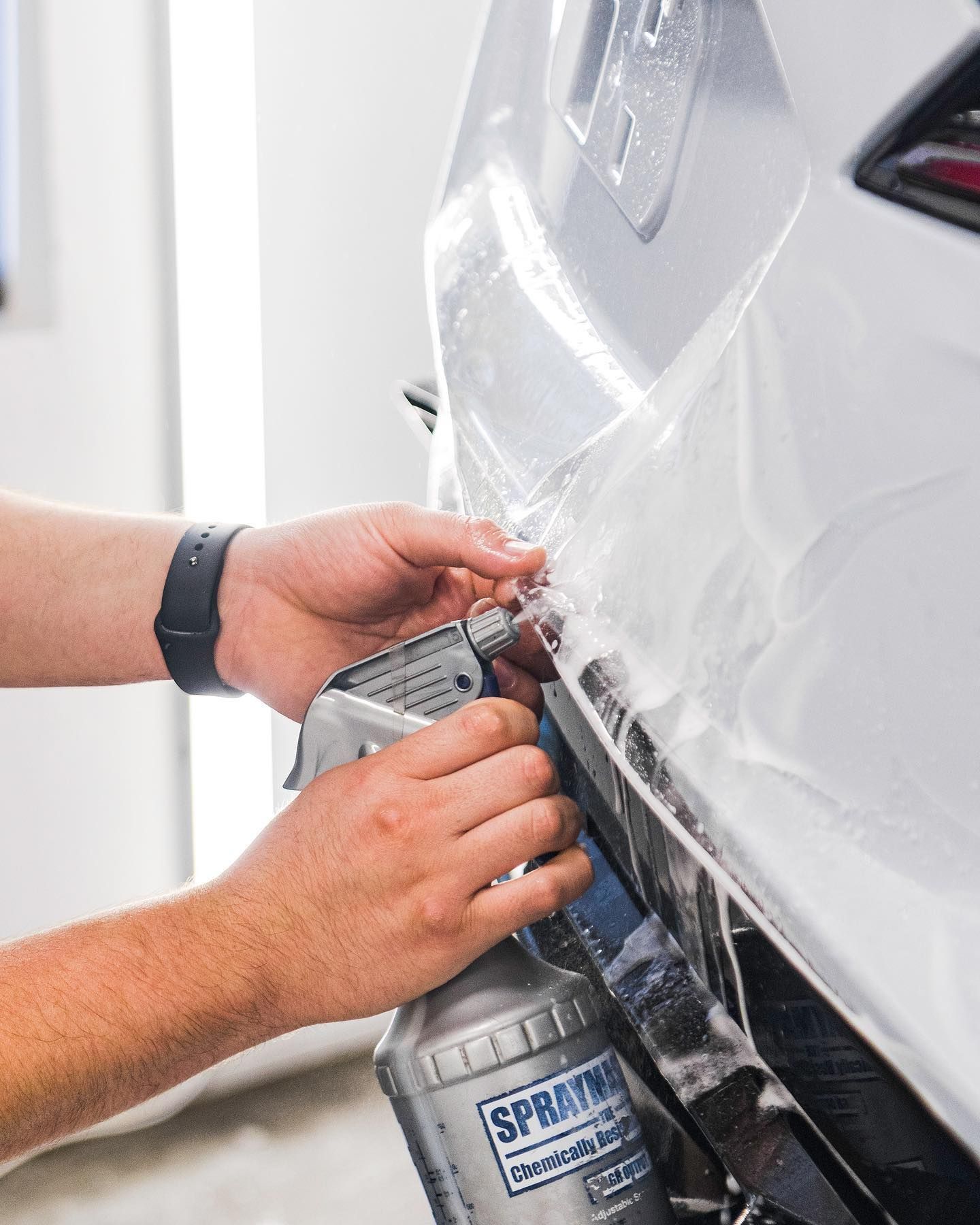 Hands applying clear protective film to a white car bumper with a spray bottle and squeegee.