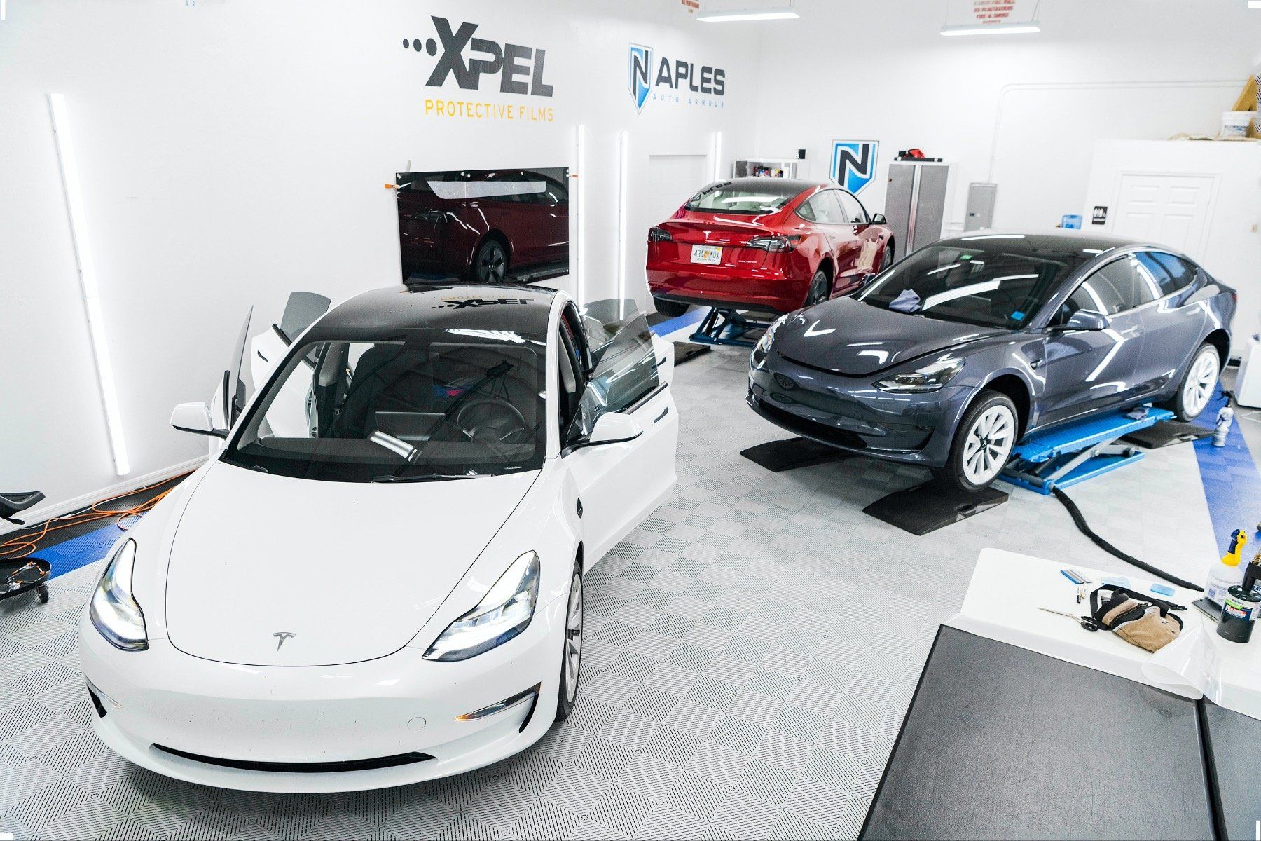 A white tesla model 3 is parked in a garage next to a gray tesla model 3.