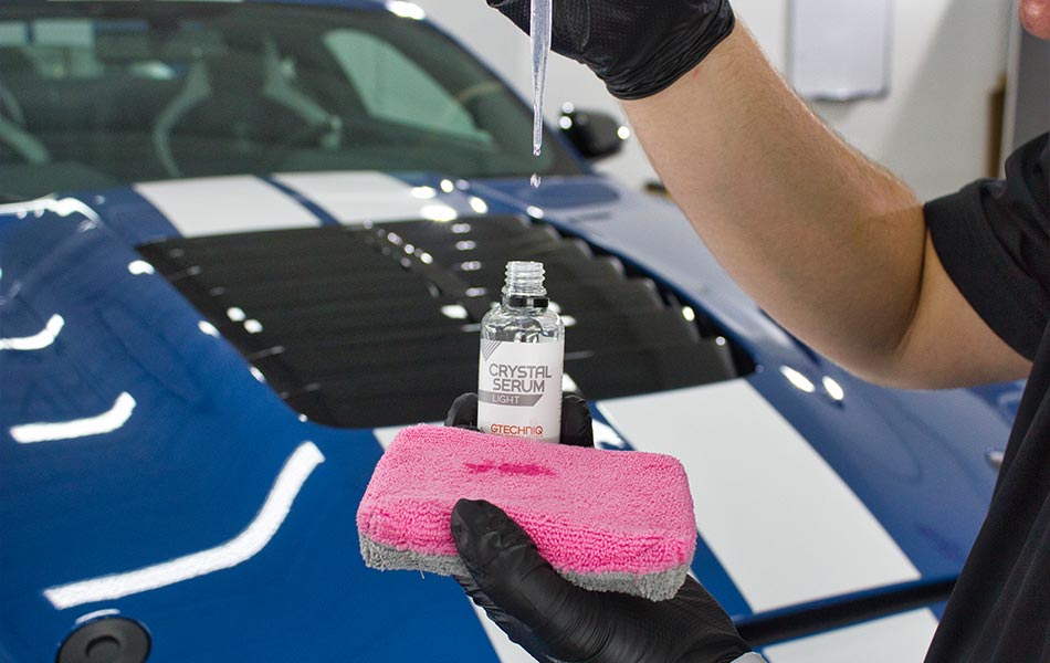 Person applying ceramic coating to a blue car's hood with a pink applicator in a workshop.