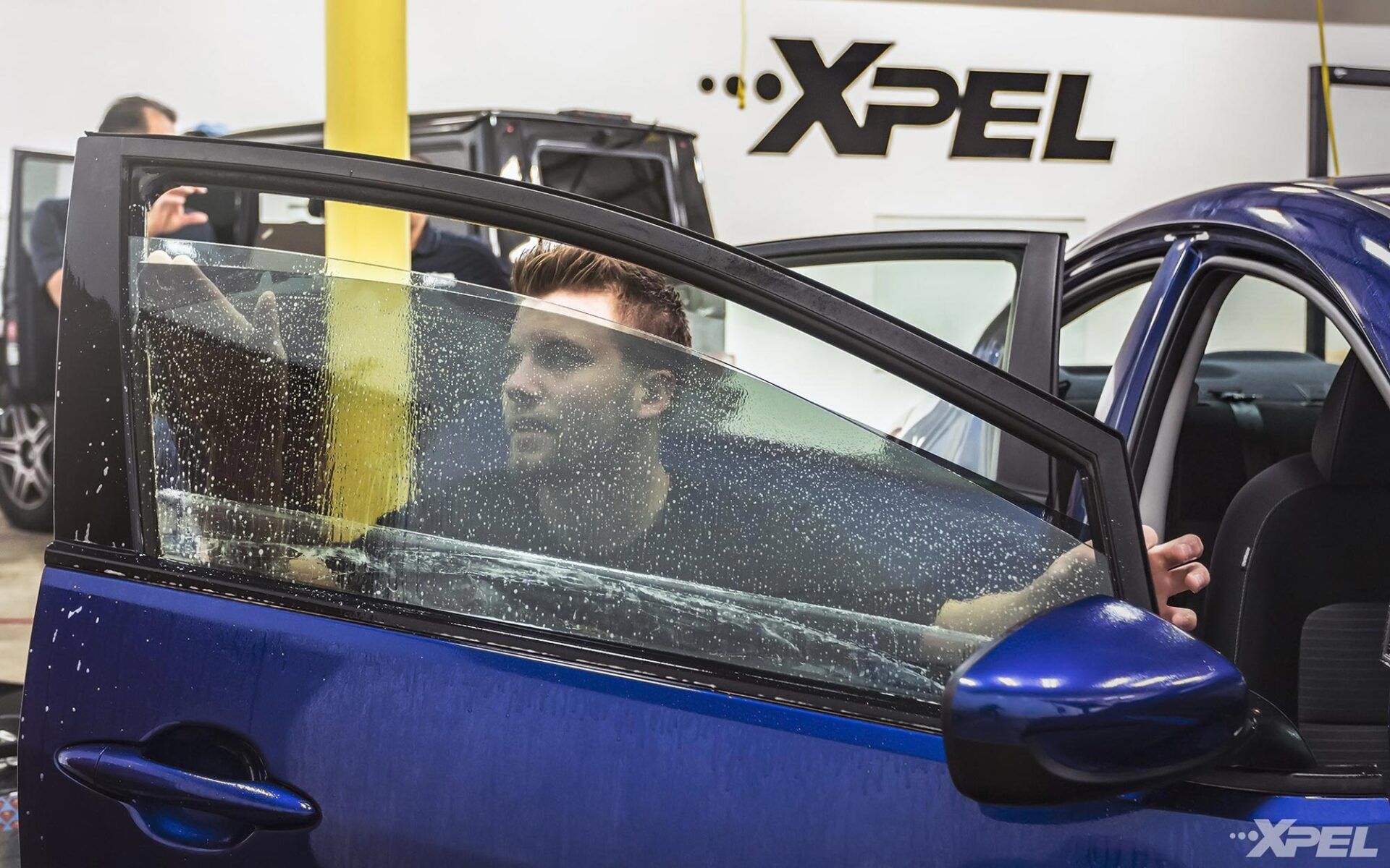 A man is sitting in a blue car with a xpel logo in the background.