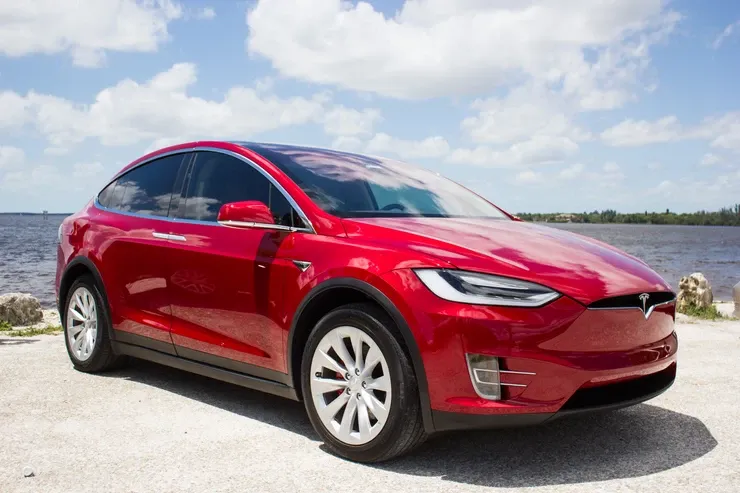A red tesla model x is parked next to a body of water.