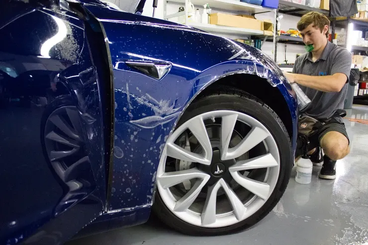 A man is kneeling down next to a blue tesla model 3 in a garage.
