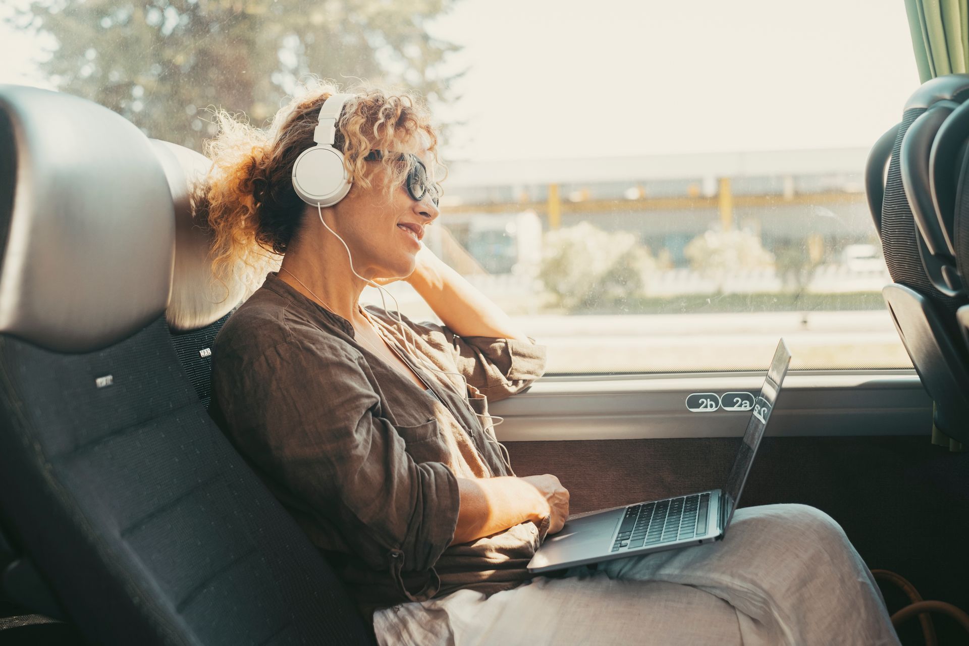 A smiling woman is listening to music with a laptop, sitting on a private bus charter seat.