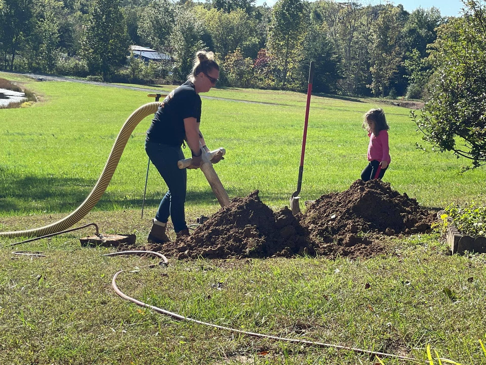 A woman is digging a hole in the ground while a little girl sits on a pile of dirt.