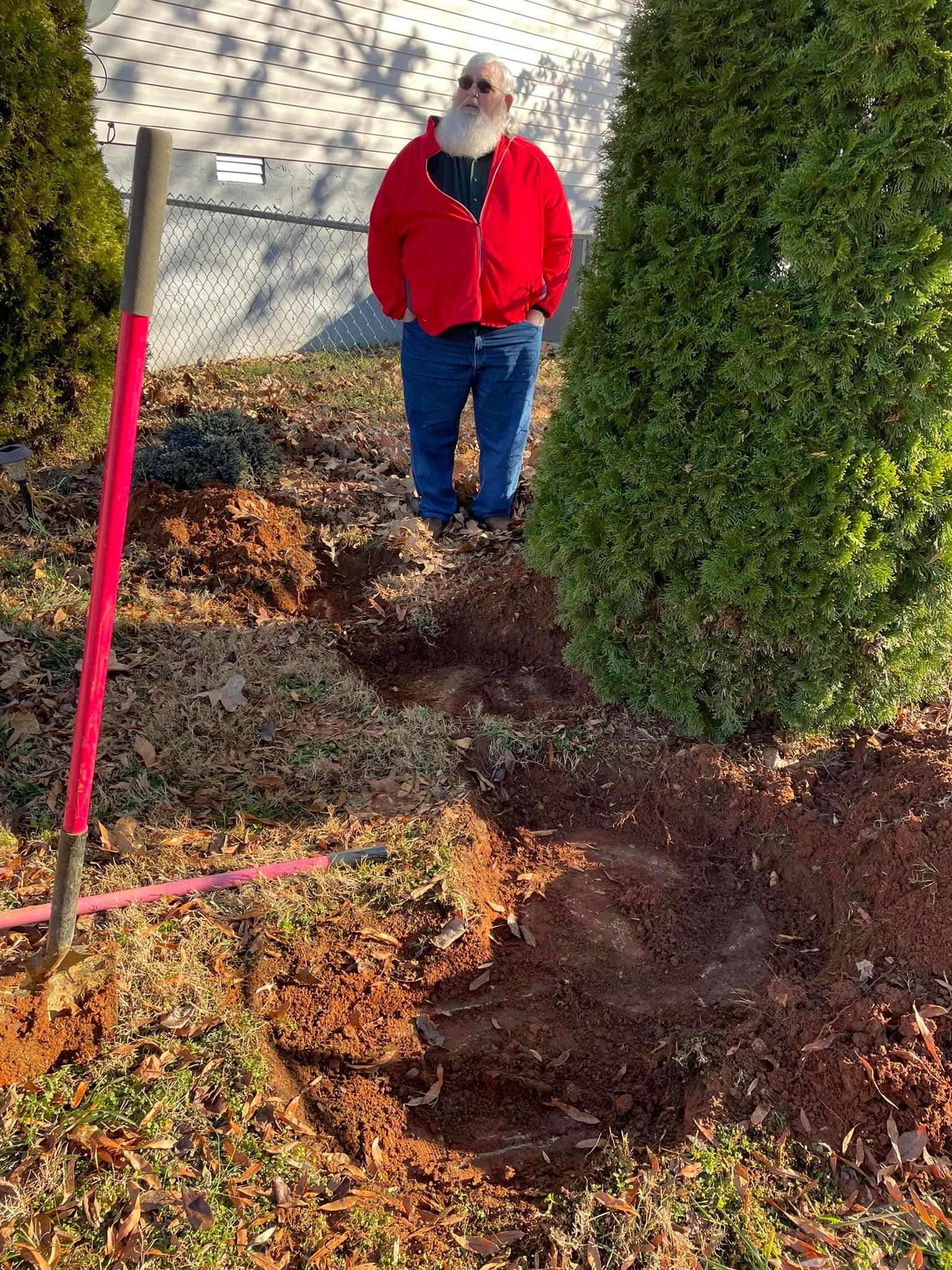 A man in a red jacket is standing in the dig hole for a septic system installation next to a tree.
