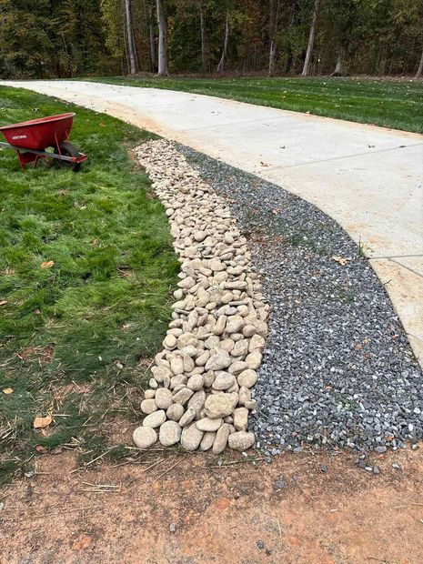 A concrete driveway edged with a strip of dark crushed stone and a wider trench filled with large, light-colored river rocks.