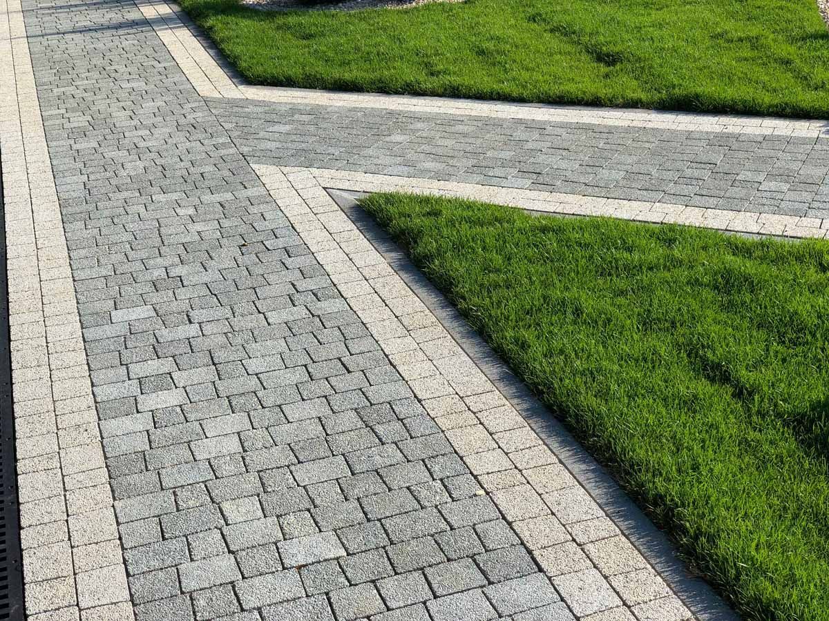 A paved garden path made of grey cobblestones with light-colored borders separating it from patches of green grass.