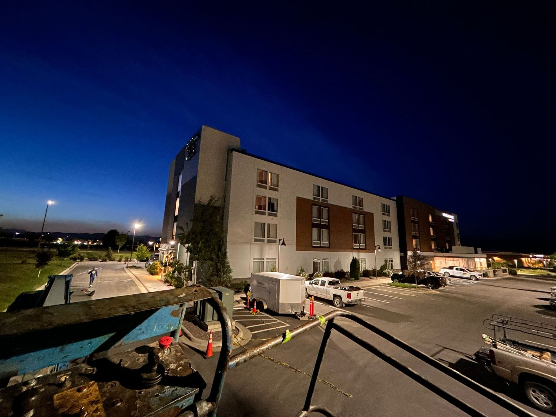 Hotel exterior at dusk with blue sky, illuminated facade, parked vehicles, and construction equipment.