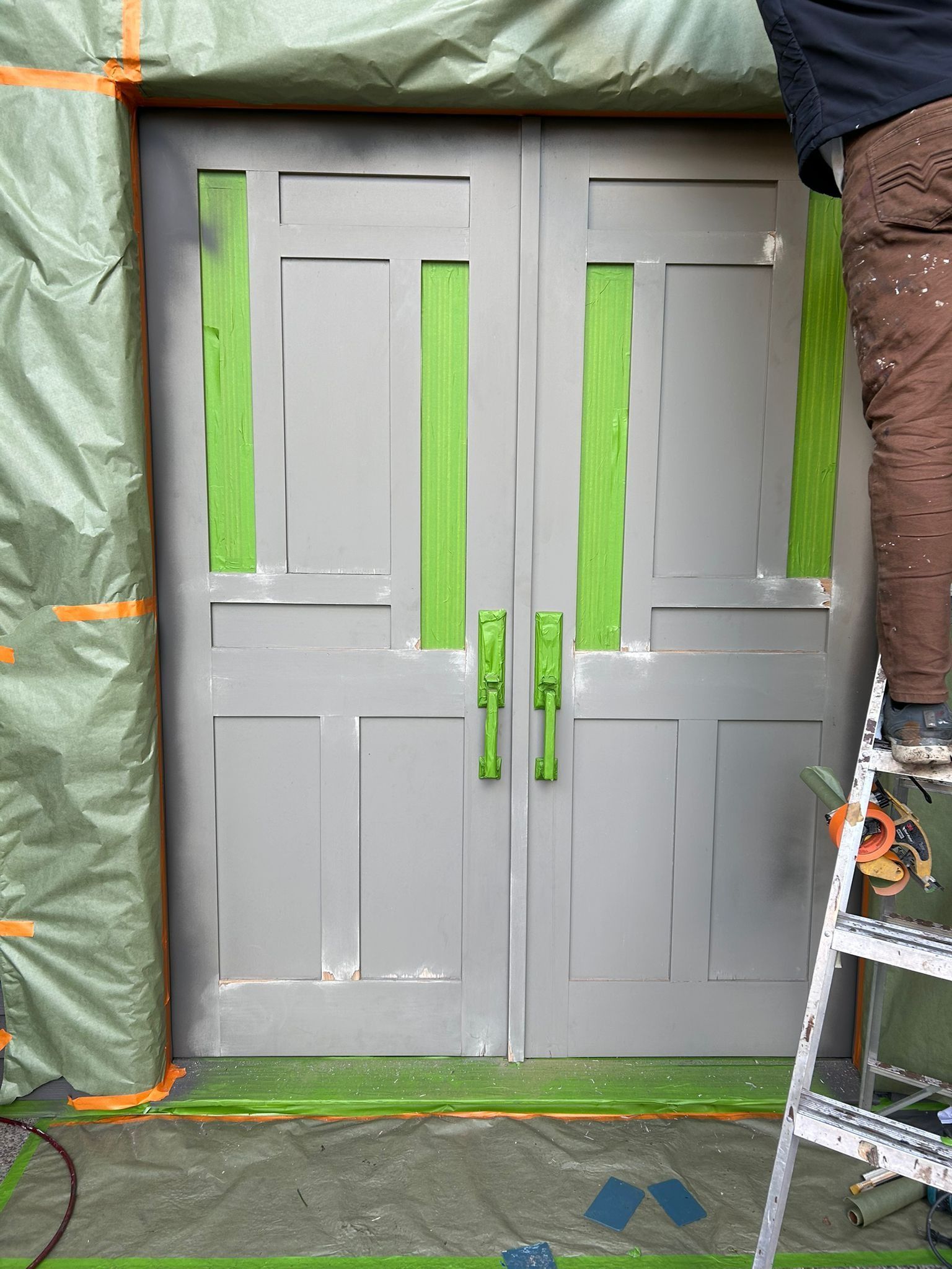 Double gray doors being painted with green handles and green tape, person on ladder.