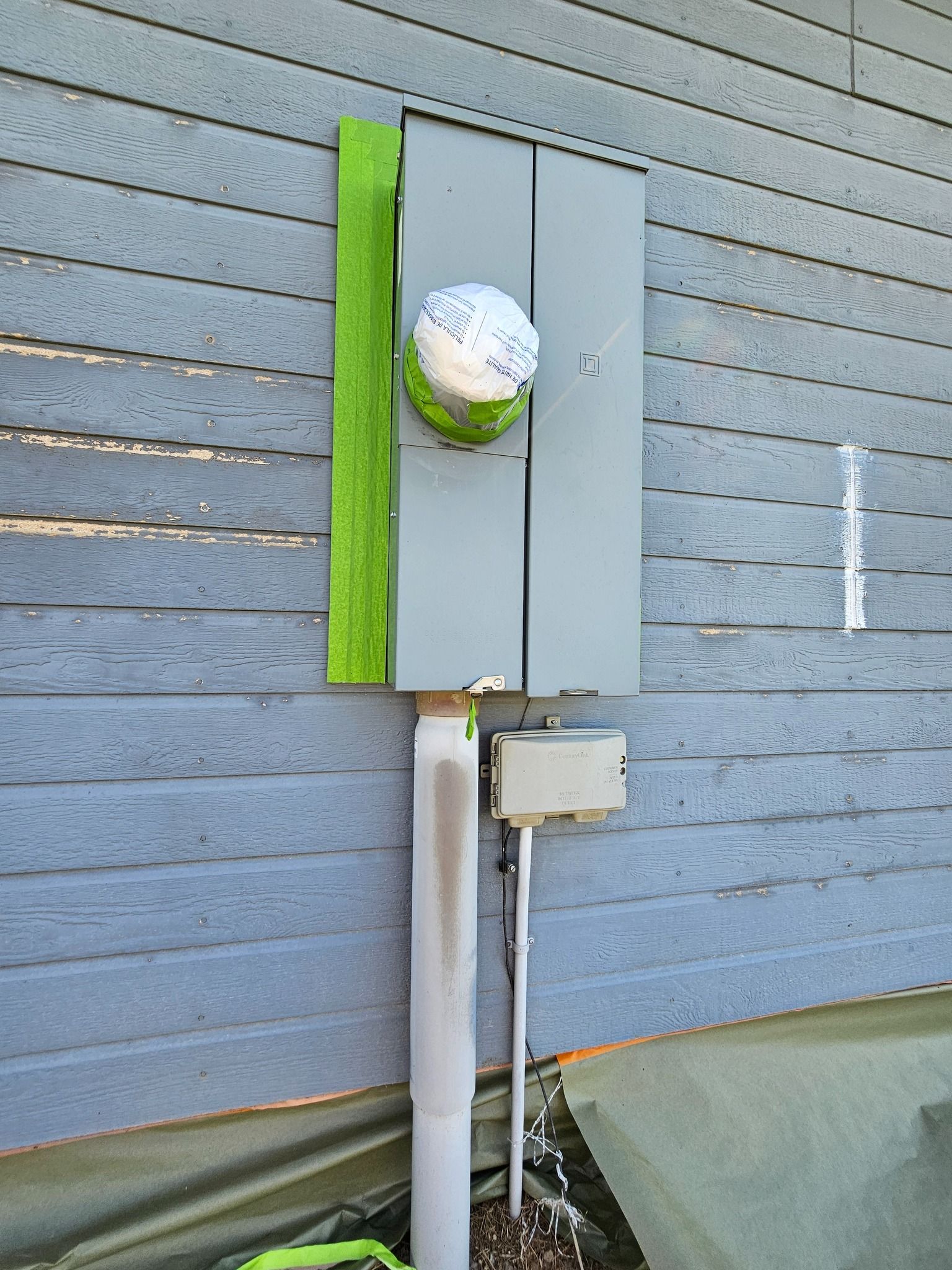 Outdoor electrical panel on a gray siding wall, with snow-covered green object and utility pipes.