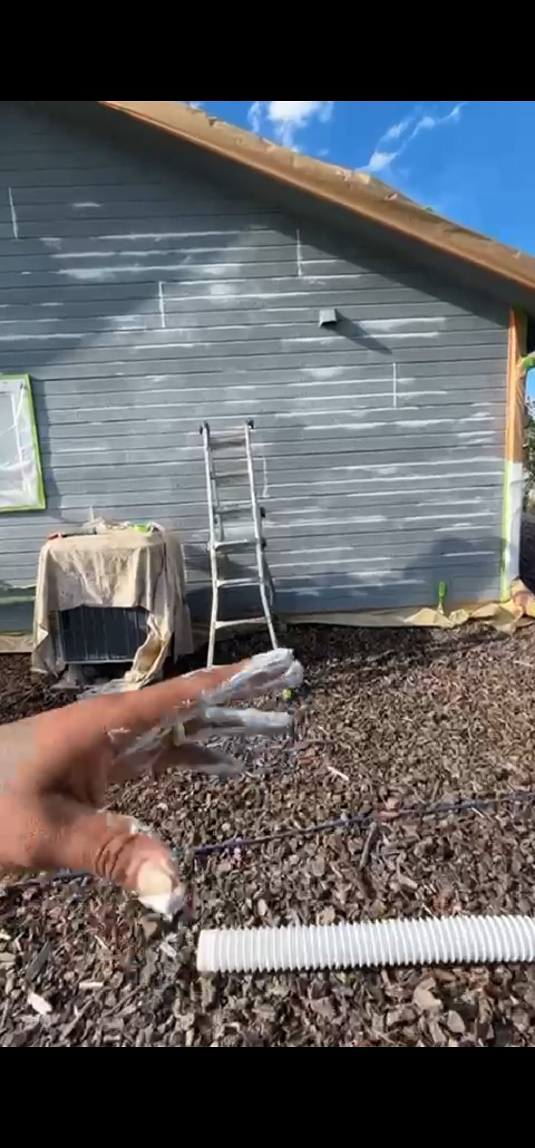Person with paint-covered fingers in front of a house being painted. Ladder and covered items are visible.
