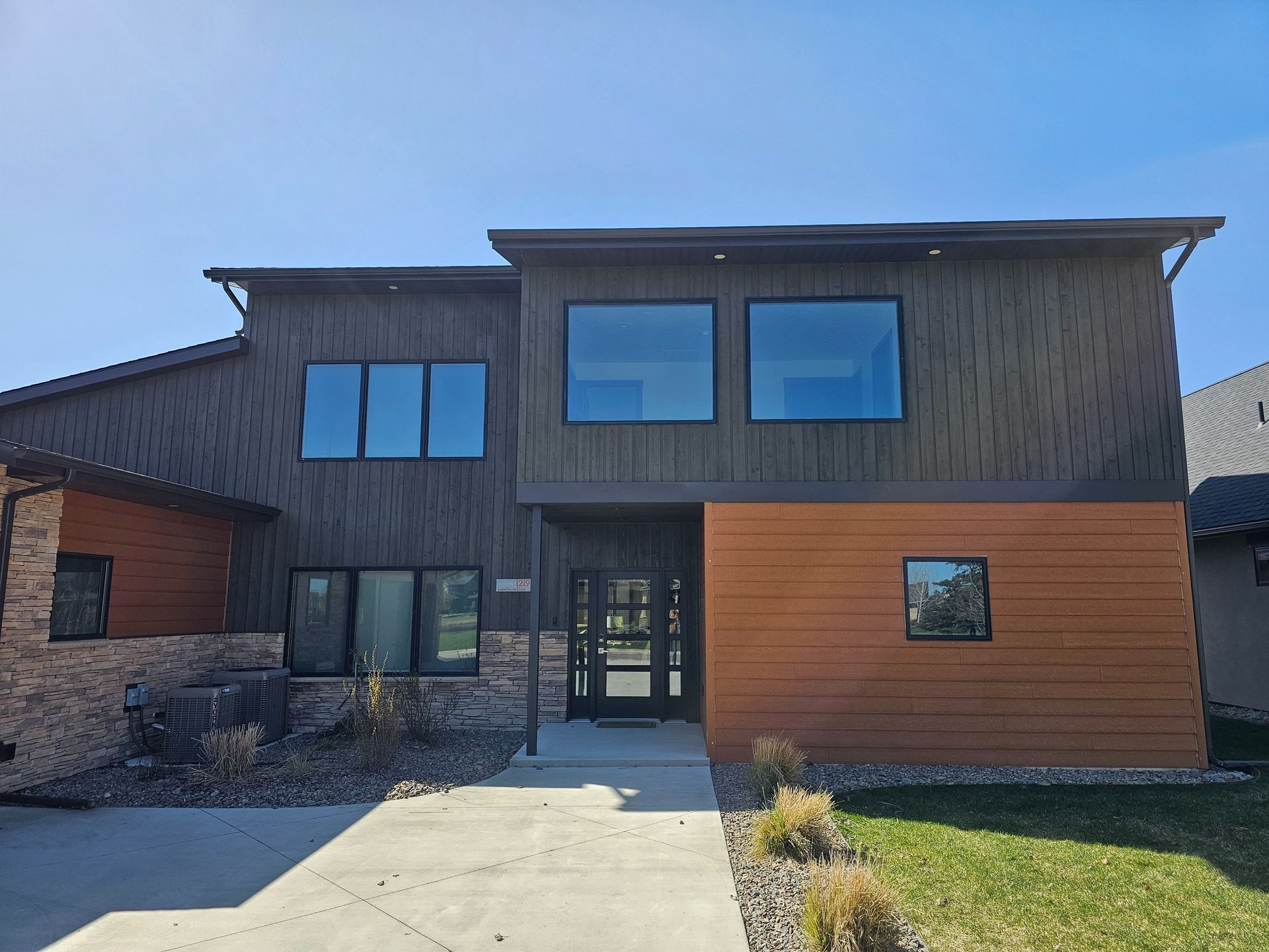 Modern two-story house with brown and orange siding, large windows, and a paved walkway.