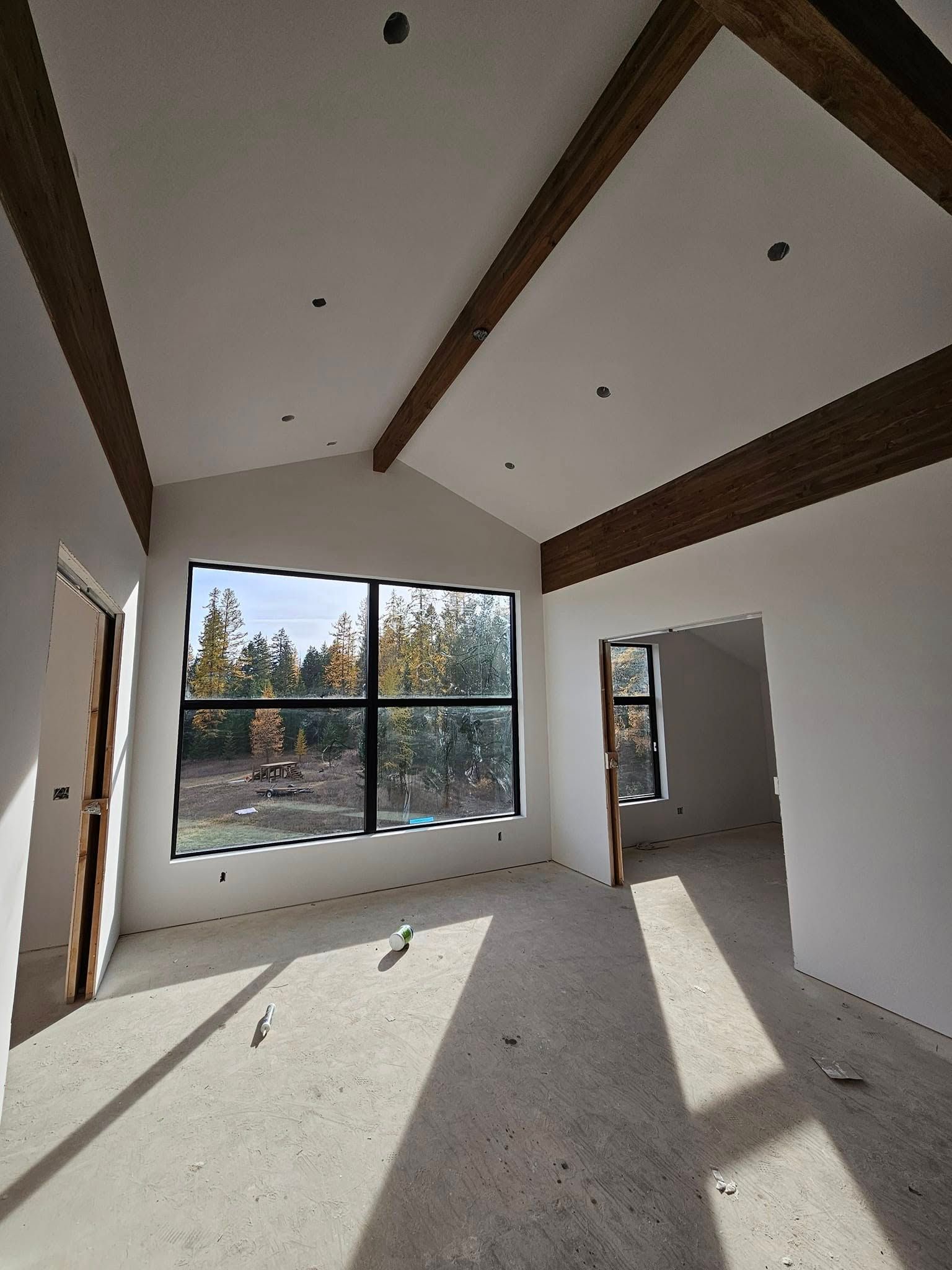 Interior of a room with large window, exposed wooden beams on white vaulted ceiling. Unfinished floor.