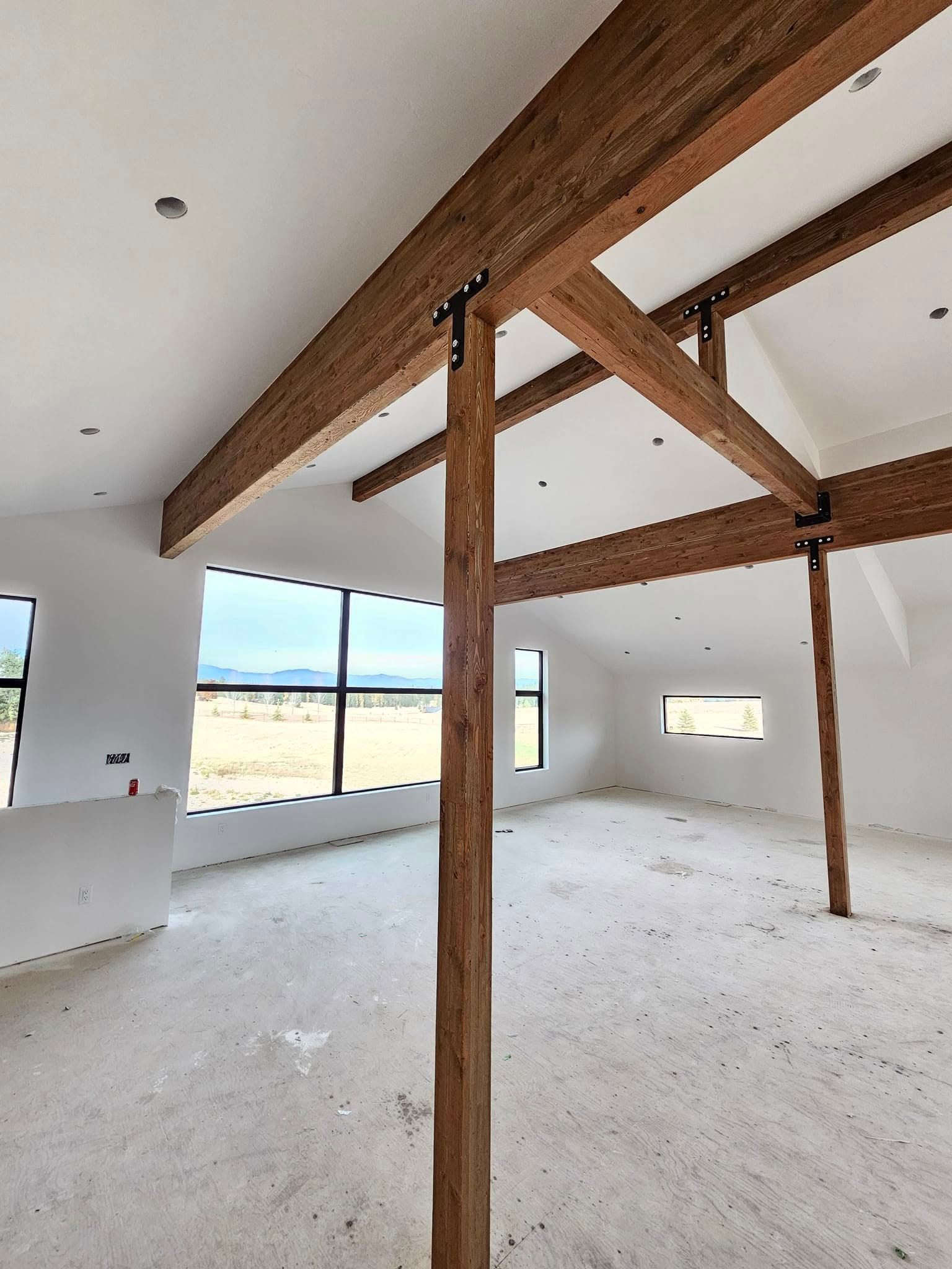 Interior view of a new home under construction with exposed wooden beams, white walls, and large windows.