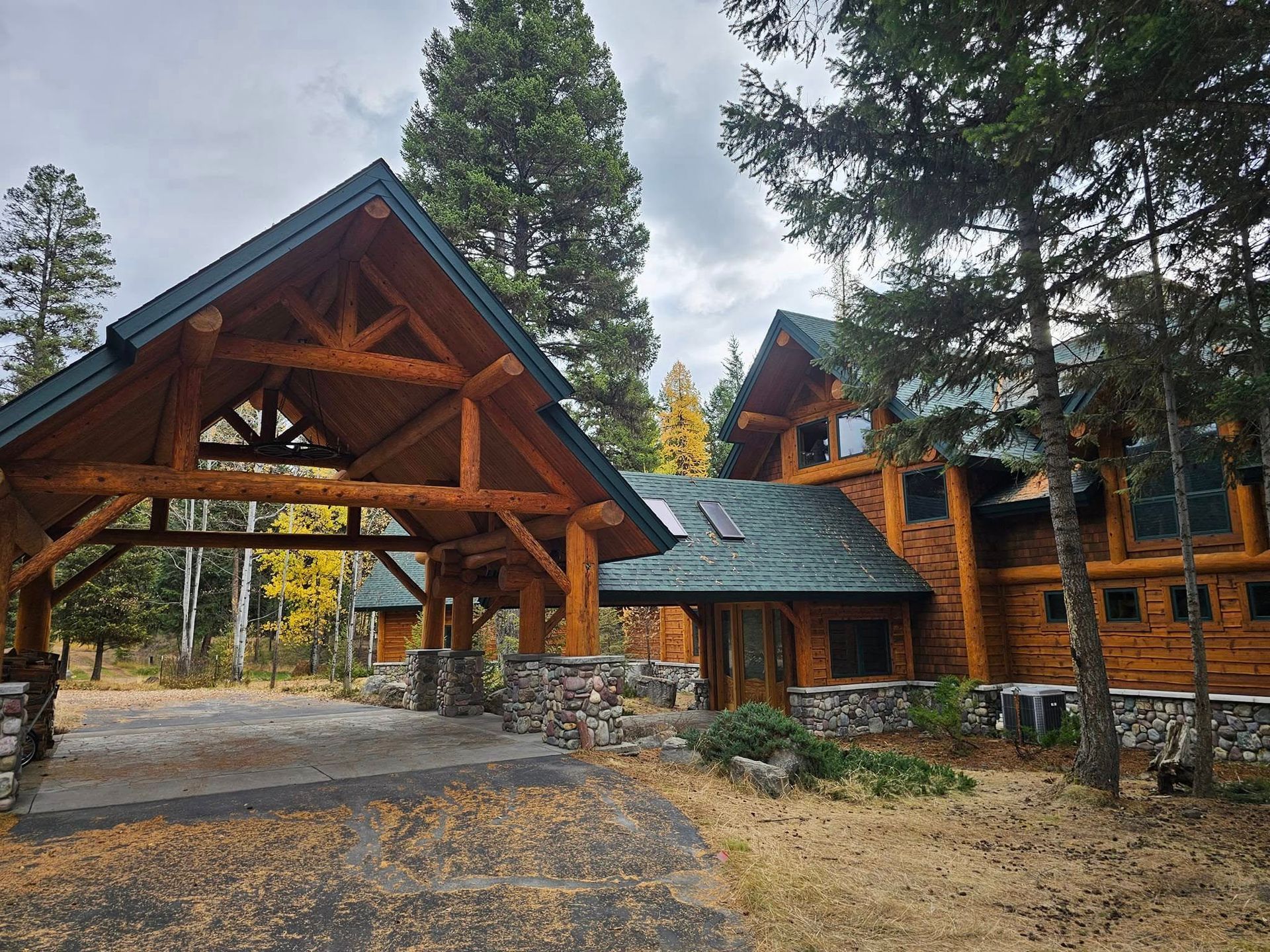Log cabin with a porte-cochere, surrounded by trees and a stone walkway under a cloudy sky.