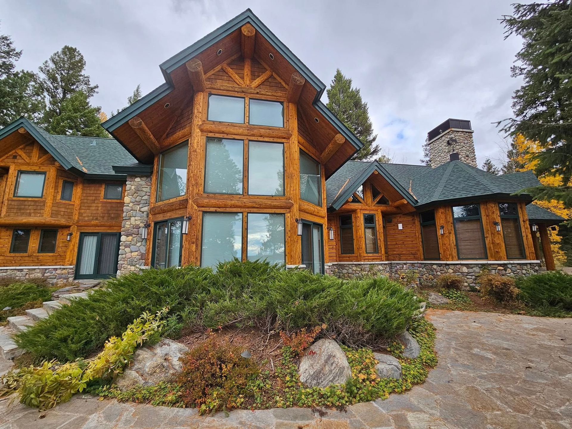 Lodge-style home with large windows, stone accents, and a stone chimney under a cloudy sky.