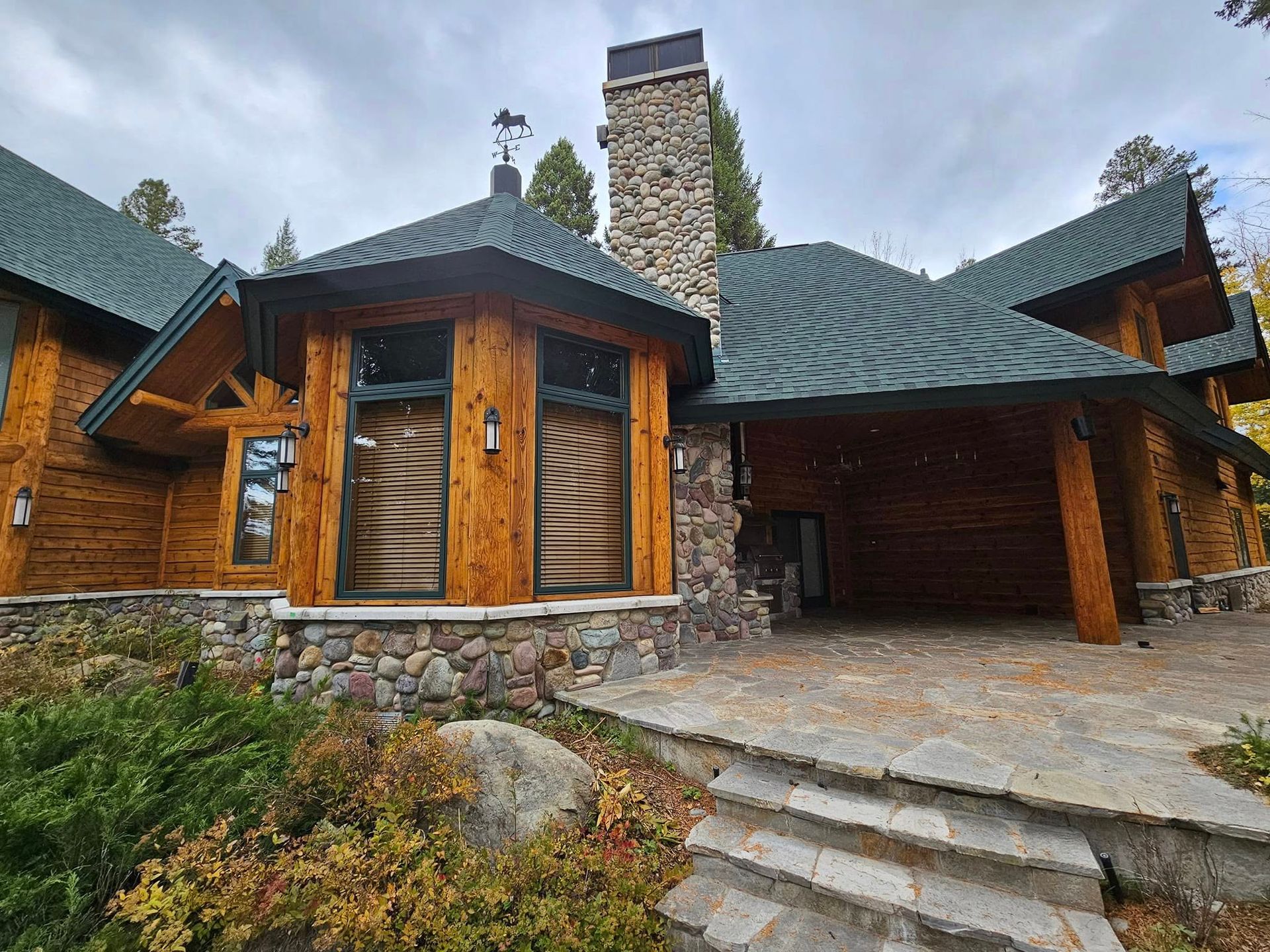 Log cabin with stone chimney and foundation; green roof; covered carport entrance.