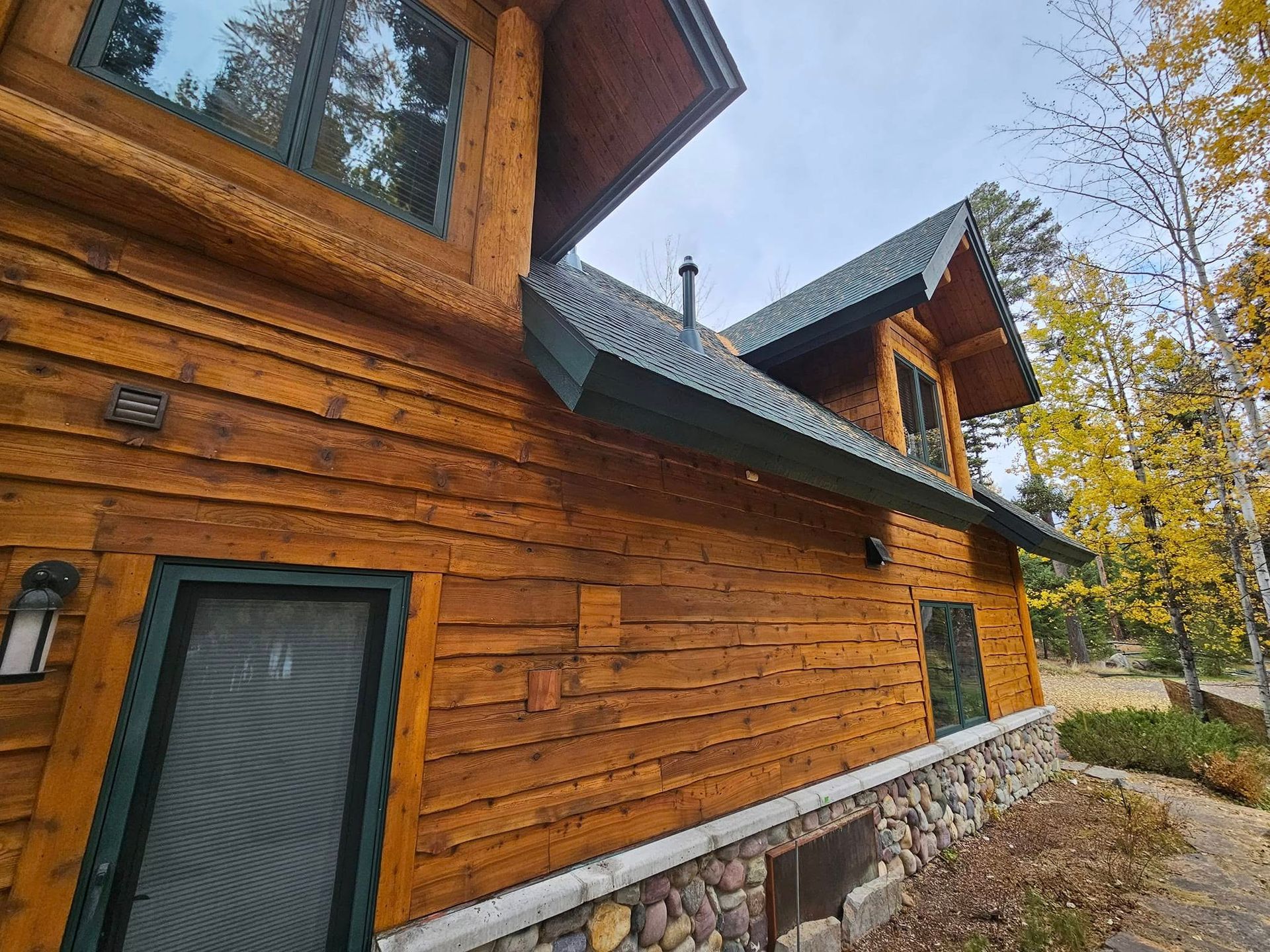 Log cabin with wood exterior, green trim, and stone foundation against a backdrop of autumn trees and sky.