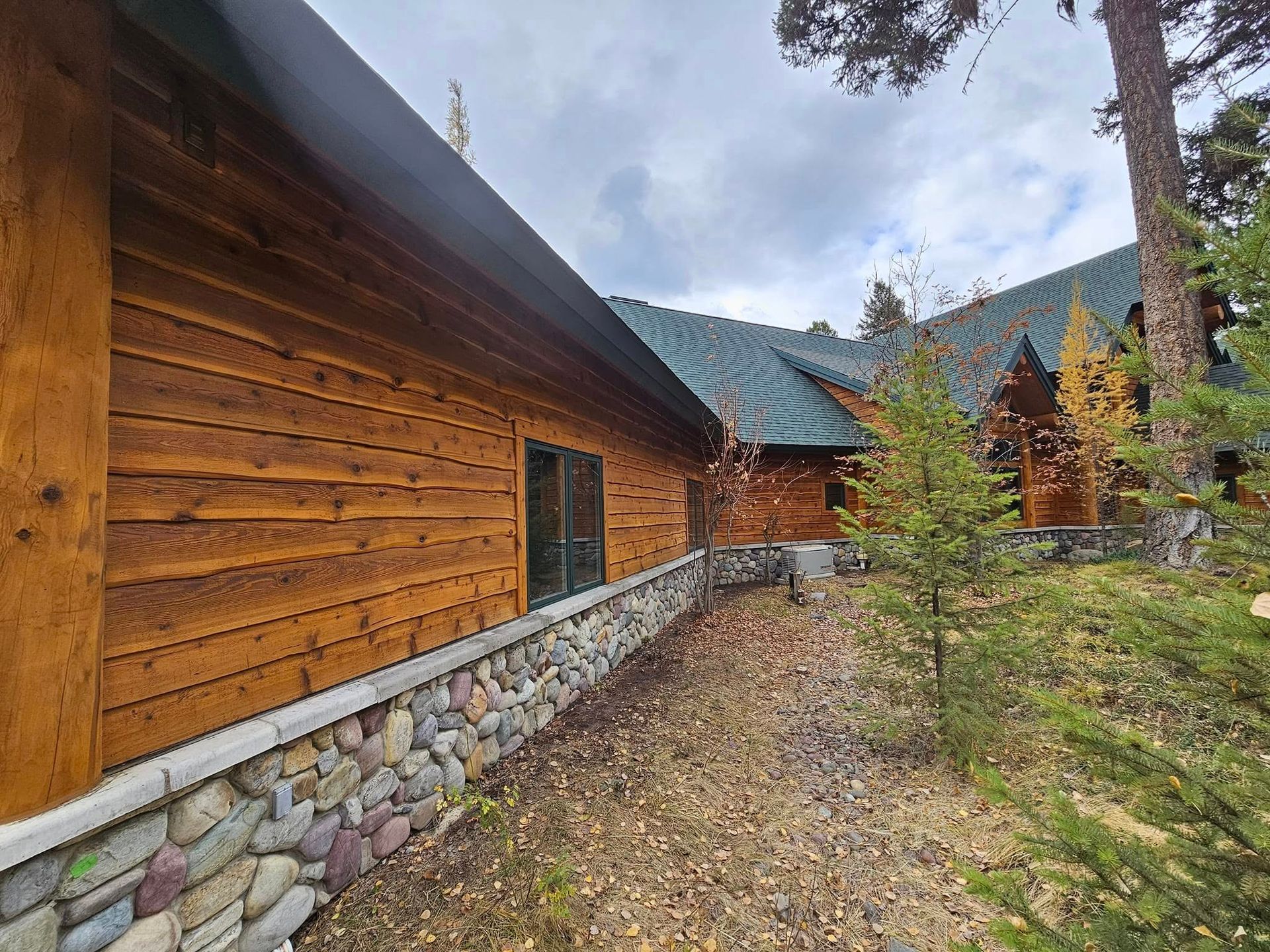 Wooden cabin exterior with stone wall, green roof, and surrounding trees under cloudy sky.