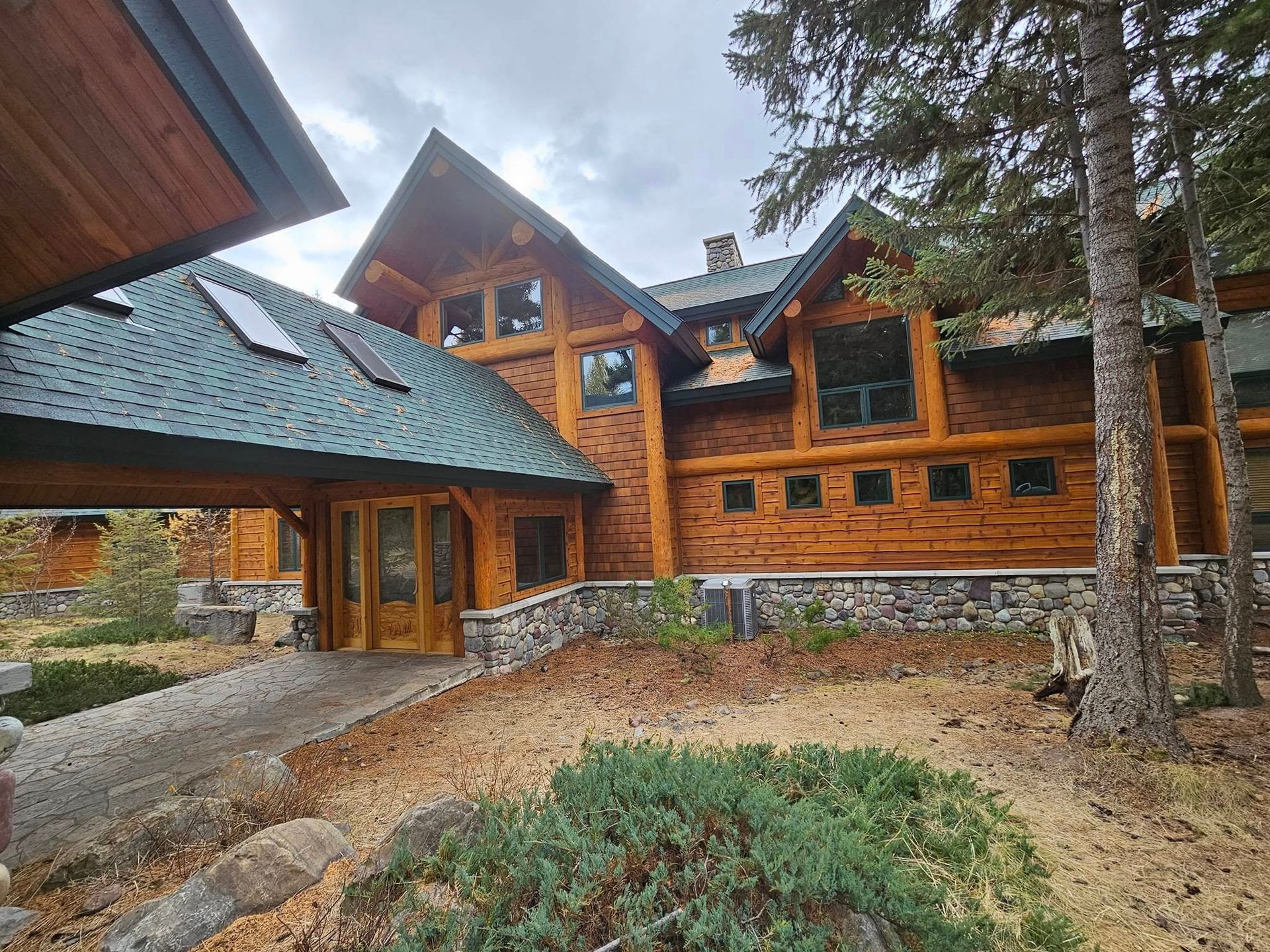 Lodge-style home with wooden logs and a green roof. A stone base and a driveway are in front.