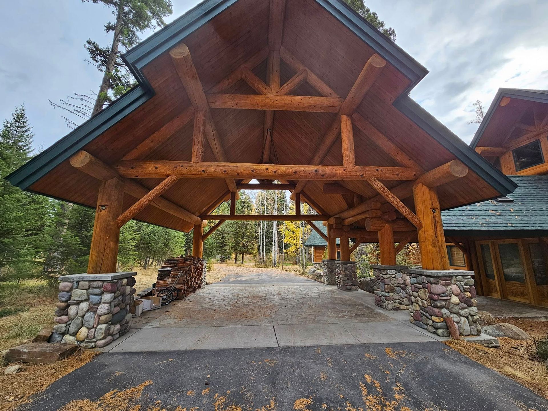 Covered driveway with wooden beams, stone pillars, and asphalt surface leading to trees.