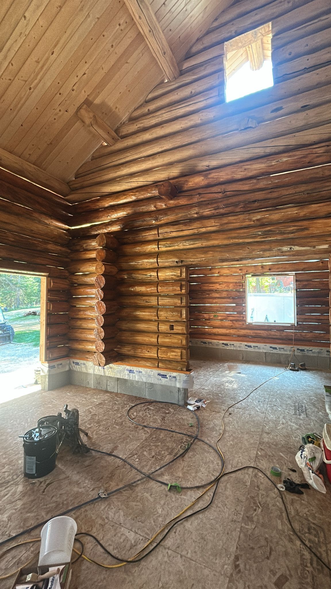 Interior of a wooden cabin under construction with logs. Walls and ceiling are made of wood; windows and doors are open.