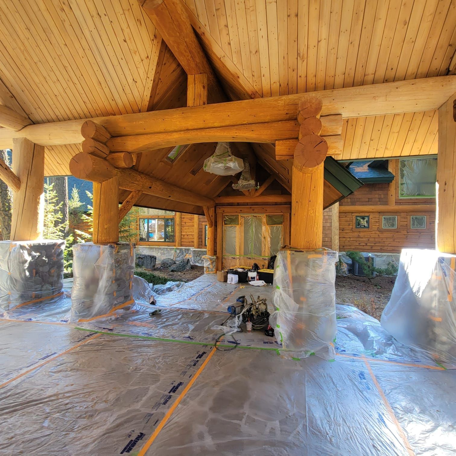 Covered porch of a log cabin with stone pillars and a wood beam structure, protective coverings on the floor.