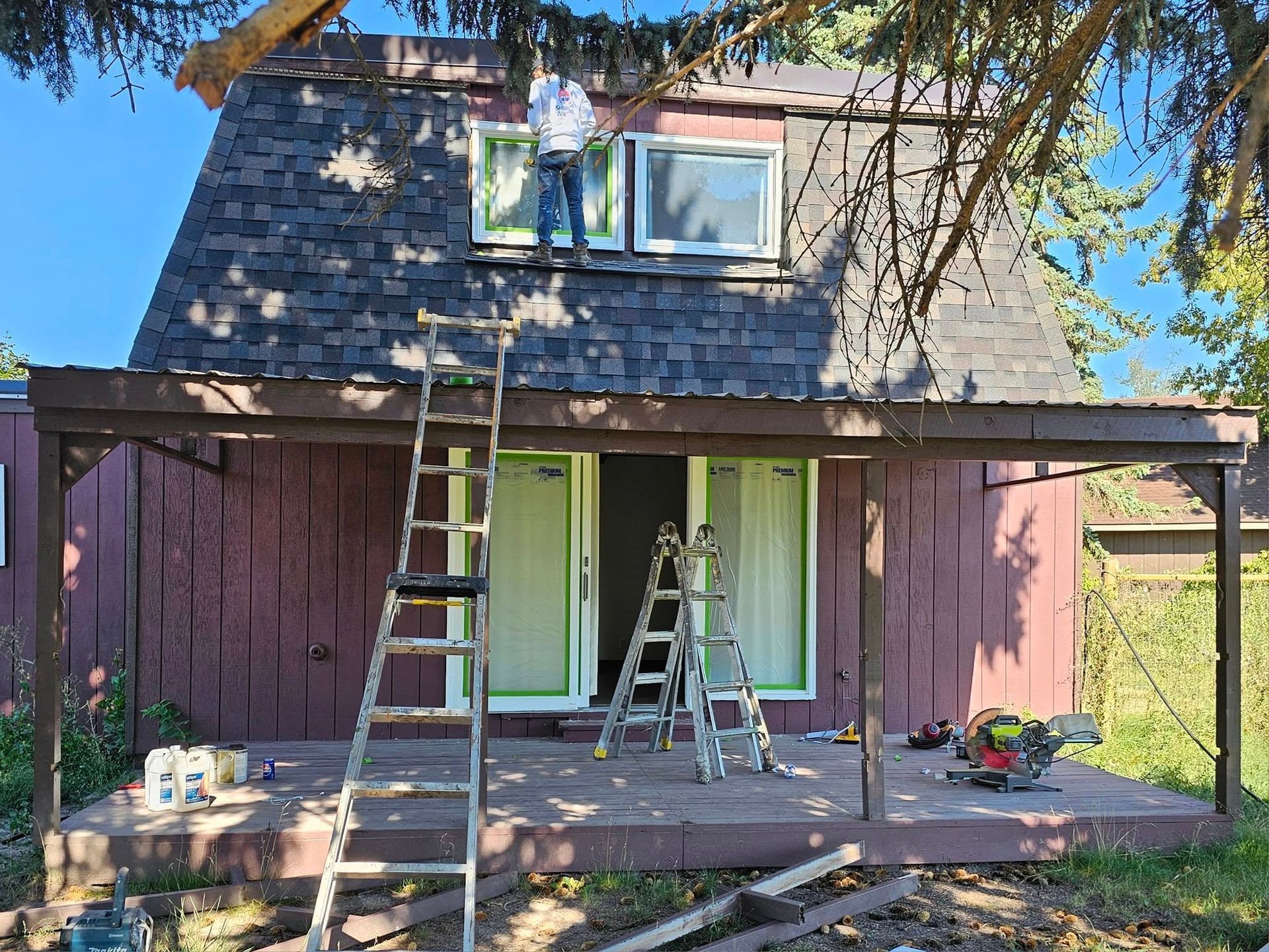 A house being painted. Person on ladder painting window trim; another on roof. Brown and red colors. Sunny day.