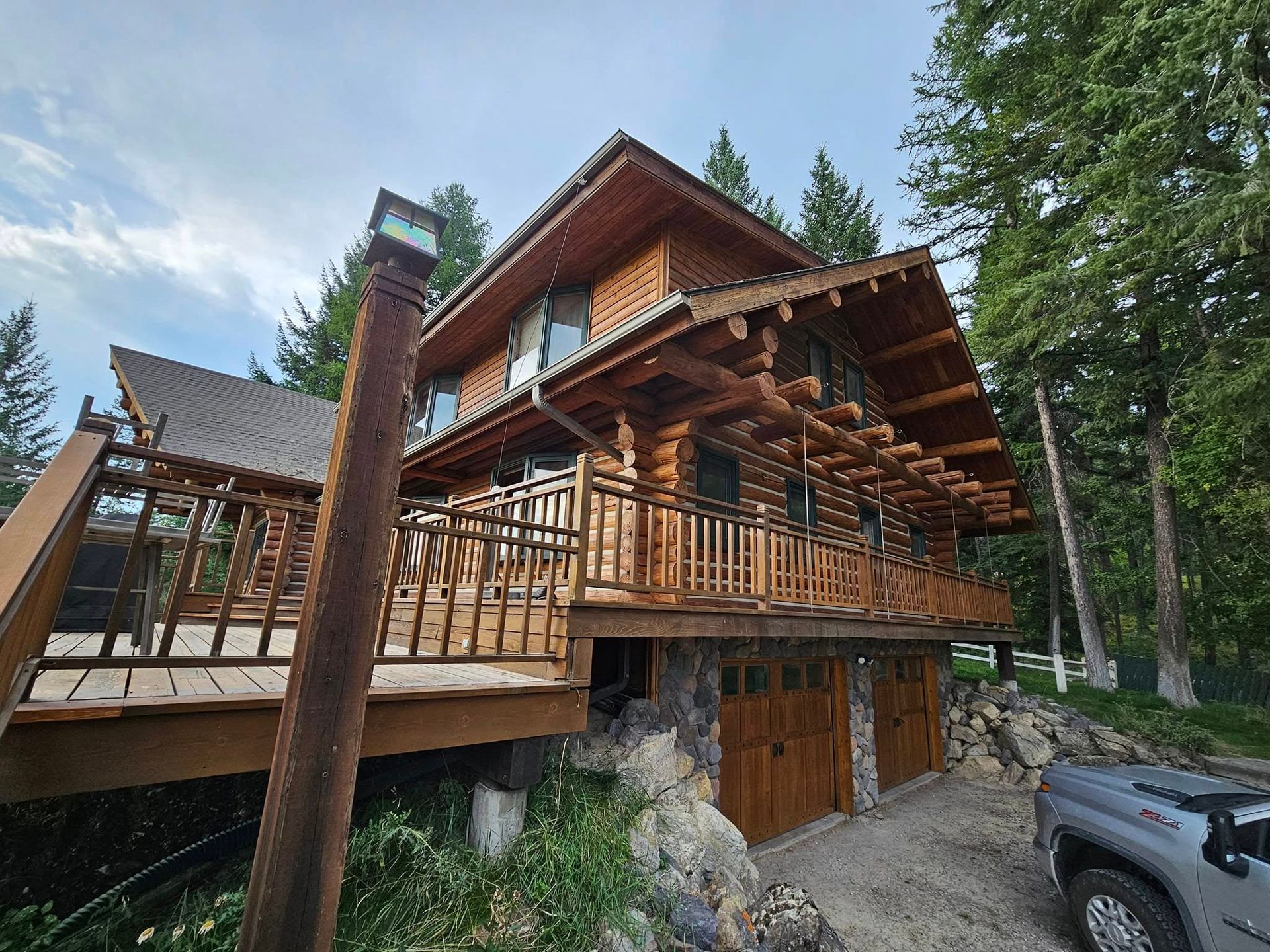 Log cabin home with a wooden deck, balcony, and attached garage. Located in a wooded area.