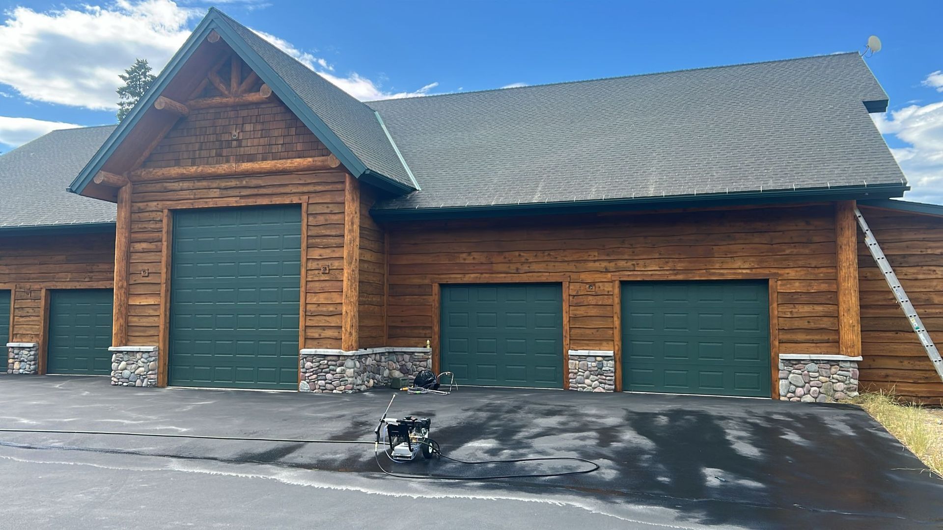 Log cabin garage with green doors, asphalt driveway, dog, and a blue sky.