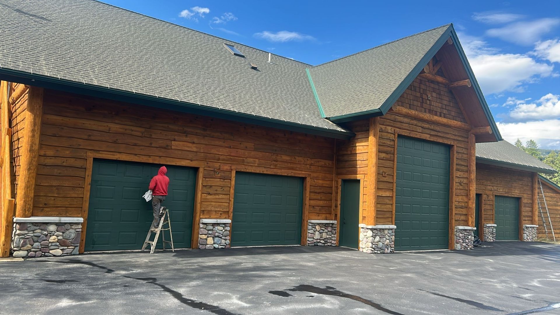 Person on ladder painting a green garage door on a log cabin-style building.