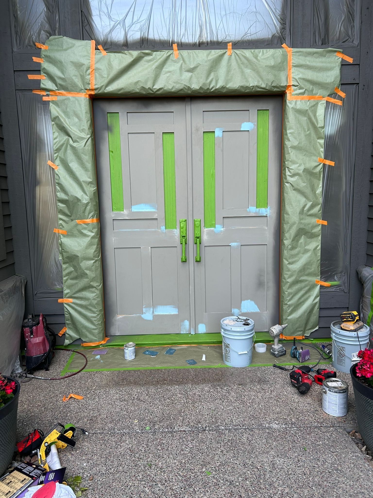 Double doors prepped for painting; green masking tape and drop cloths. Tools and paint cans on the ground.