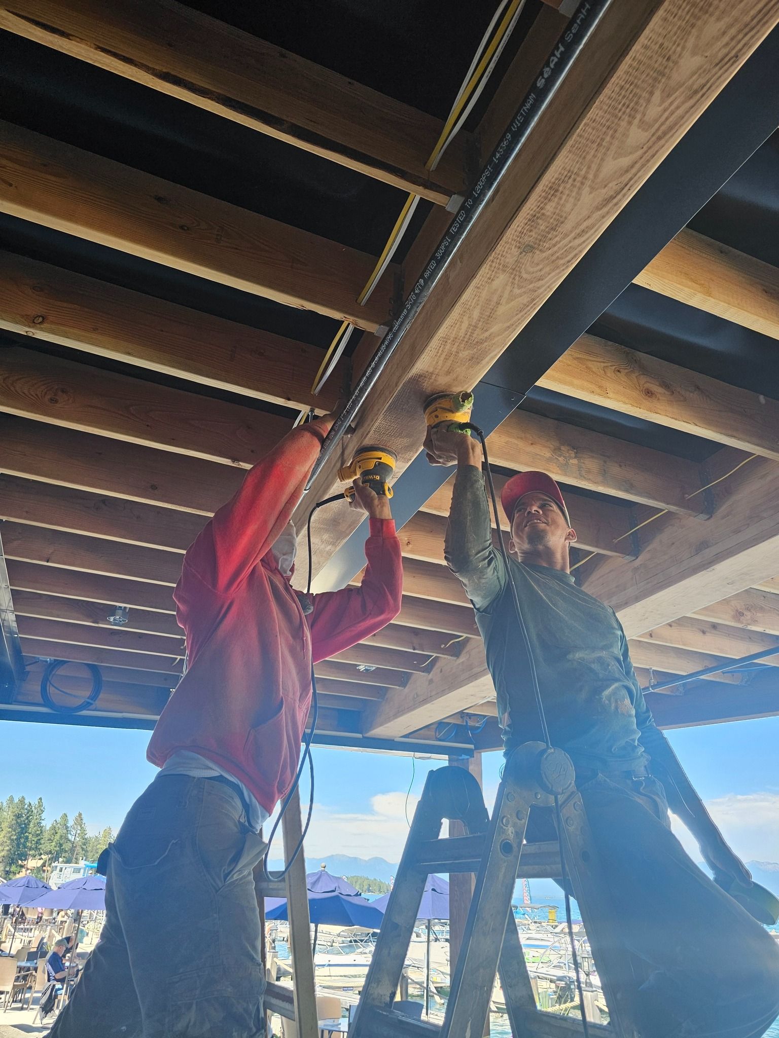 Two people on ladders drilling into a wooden beam, outdoors.
