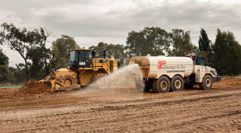 A bulldozer and a tanker truck are spraying water on a dirt field.