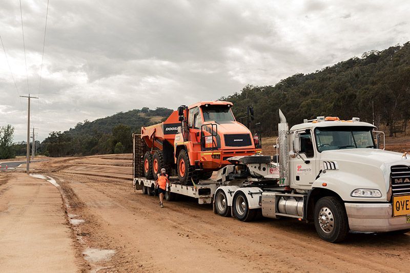 A white semi truck is carrying a large orange dump truck on a trailer.
