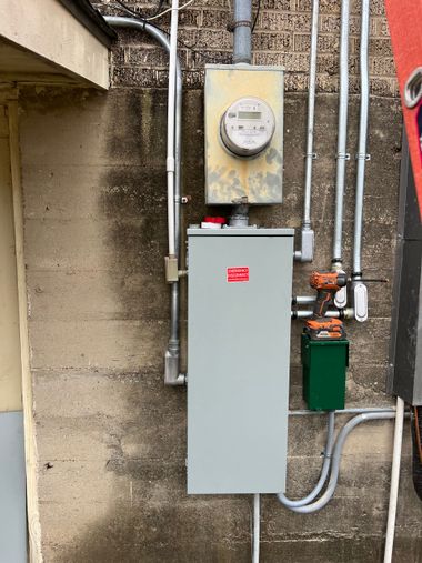 Exterior electrical meter and breaker panel mounted on a concrete wall with conduit and green utility box